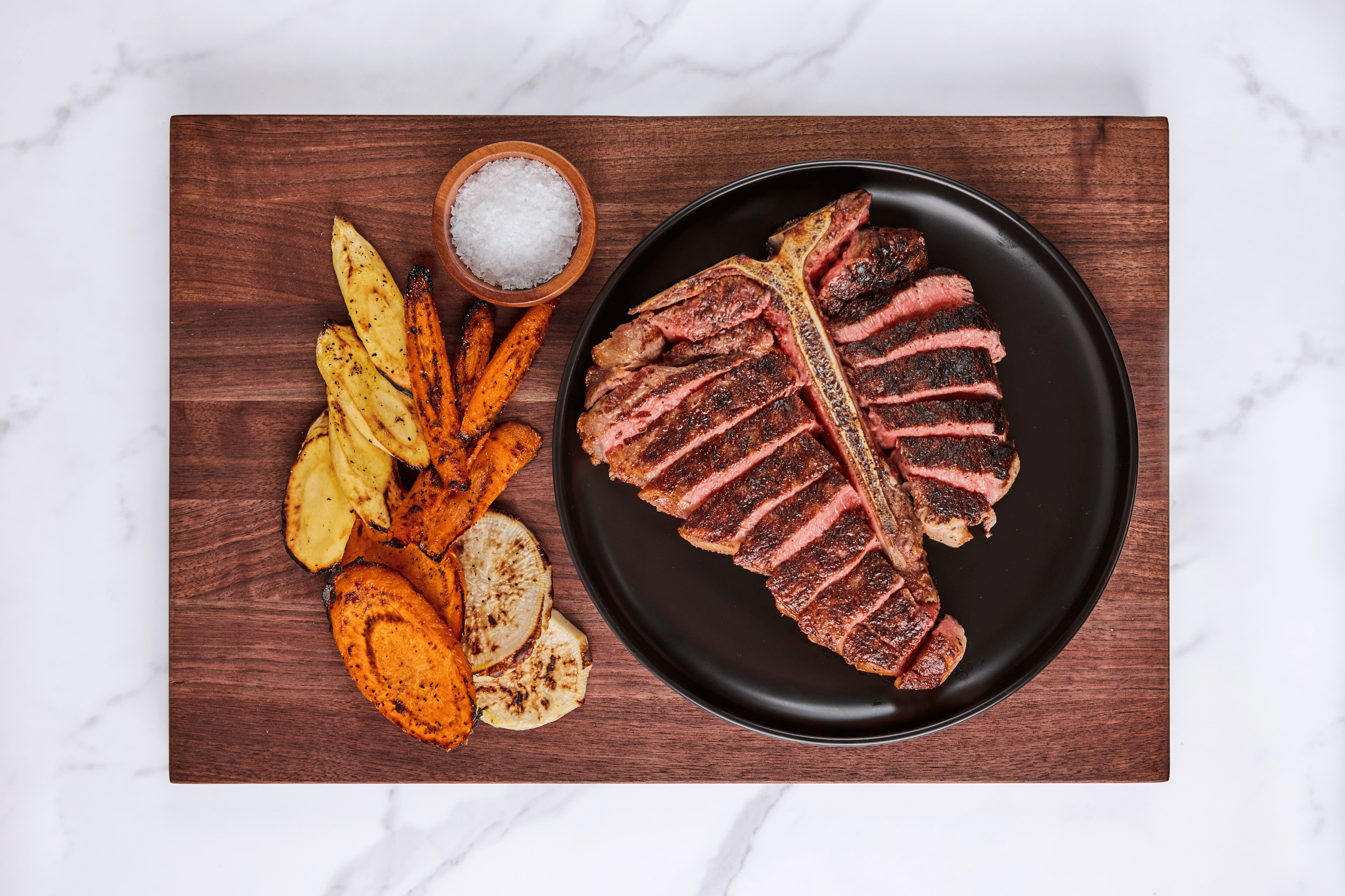 A sliced bone-in porterhouse steak on a dark plate served on a wooden board with roasted vegetables and sea salt. A sliced bone-in porterhouse steak on a dark plate served on a wooden board with roasted vegetables and sea salt.