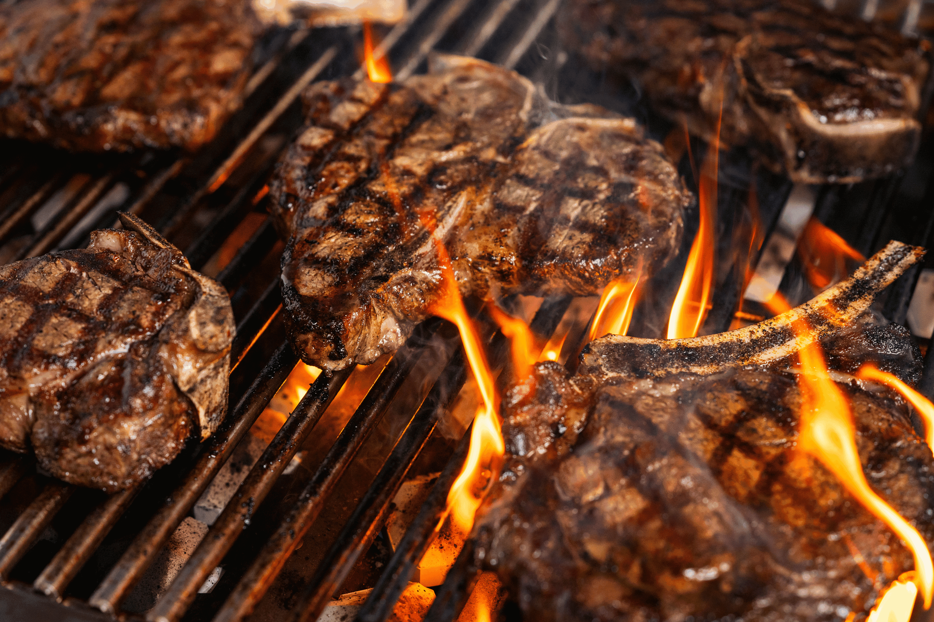 Thick bone-in ribeye steaks with dark grill marks and a heavy sear cooking over roaring open flames on a charcoal grill. Thick bone-in ribeye steaks with dark grill marks and a heavy sear cooking over roaring open flames on a charcoal grill.