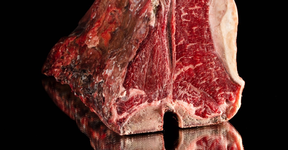 A dry-aged beef cut with a visible pellicle crust displayed upright against a black background with a mirror reflection.