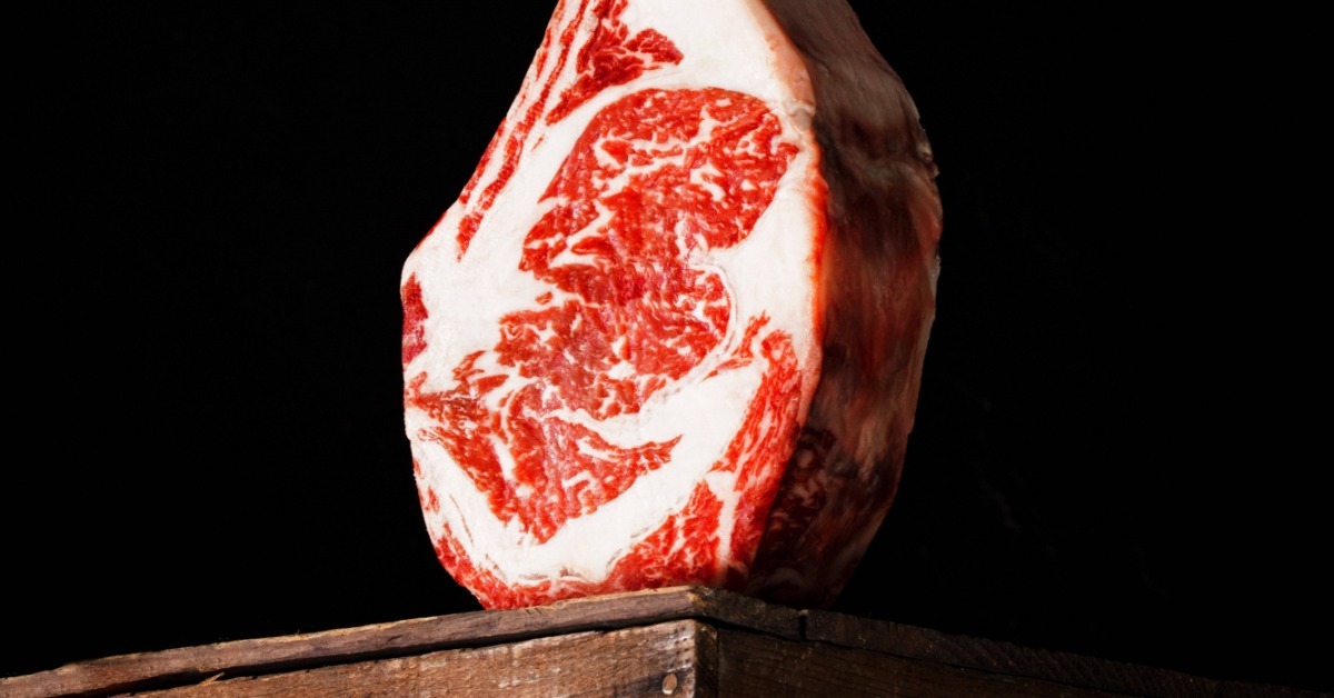 A heavily marbled raw tomahawk steak standing upright on a wooden block against a dark black background.