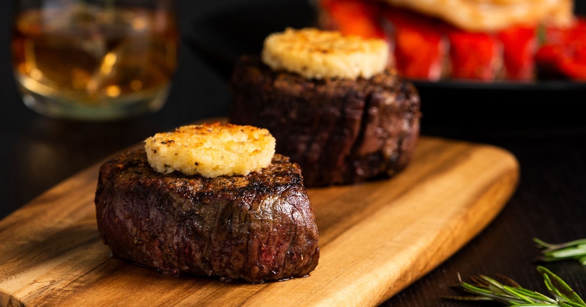 Two seared filet mignon cuts topped with compound butter on a wooden board with rosemary and a whiskey glass nearby.