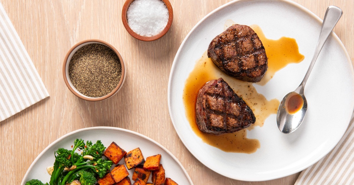 Two grilled filet mignon steaks on a white plate with pan juices beside roasted sweet potatoes and broccolini with salt and pepper.