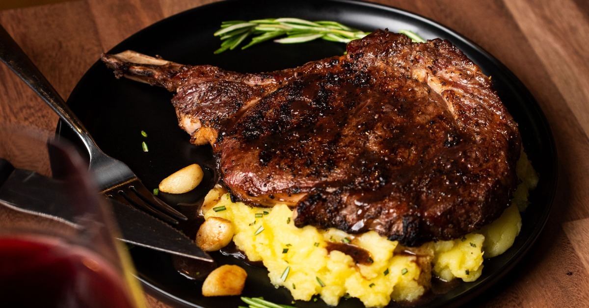 A bone-in ribeye with a charred crust served over mashed potatoes with roasted garlic and fresh rosemary on a dark plate.