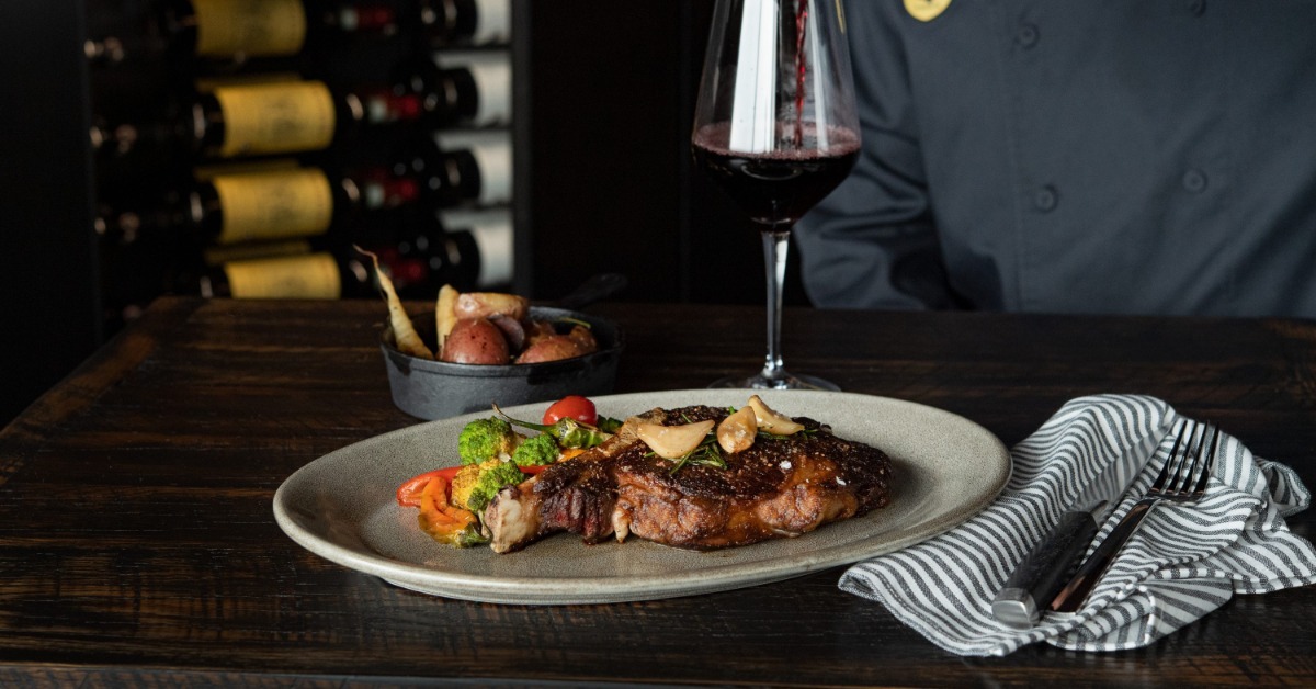 A plated seared ribeye with colorful roasted vegetables beside a glass of red wine being poured in a dimly lit dining setting.
