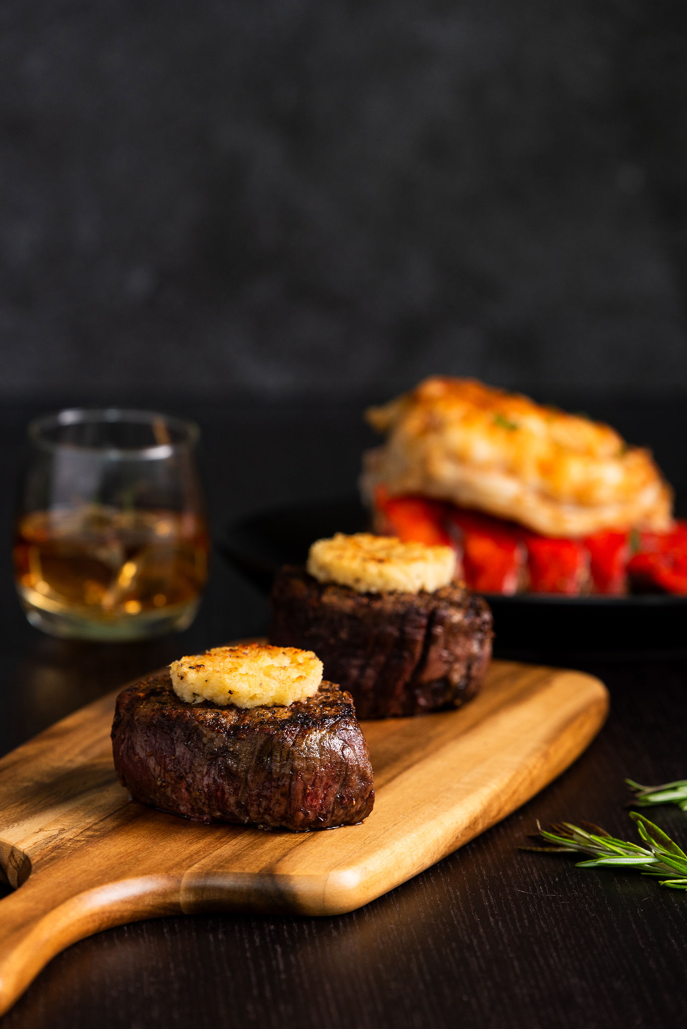 Two seared filet mignon steaks topped with compound butter on a wooden board, with whiskey and a plate of sides.