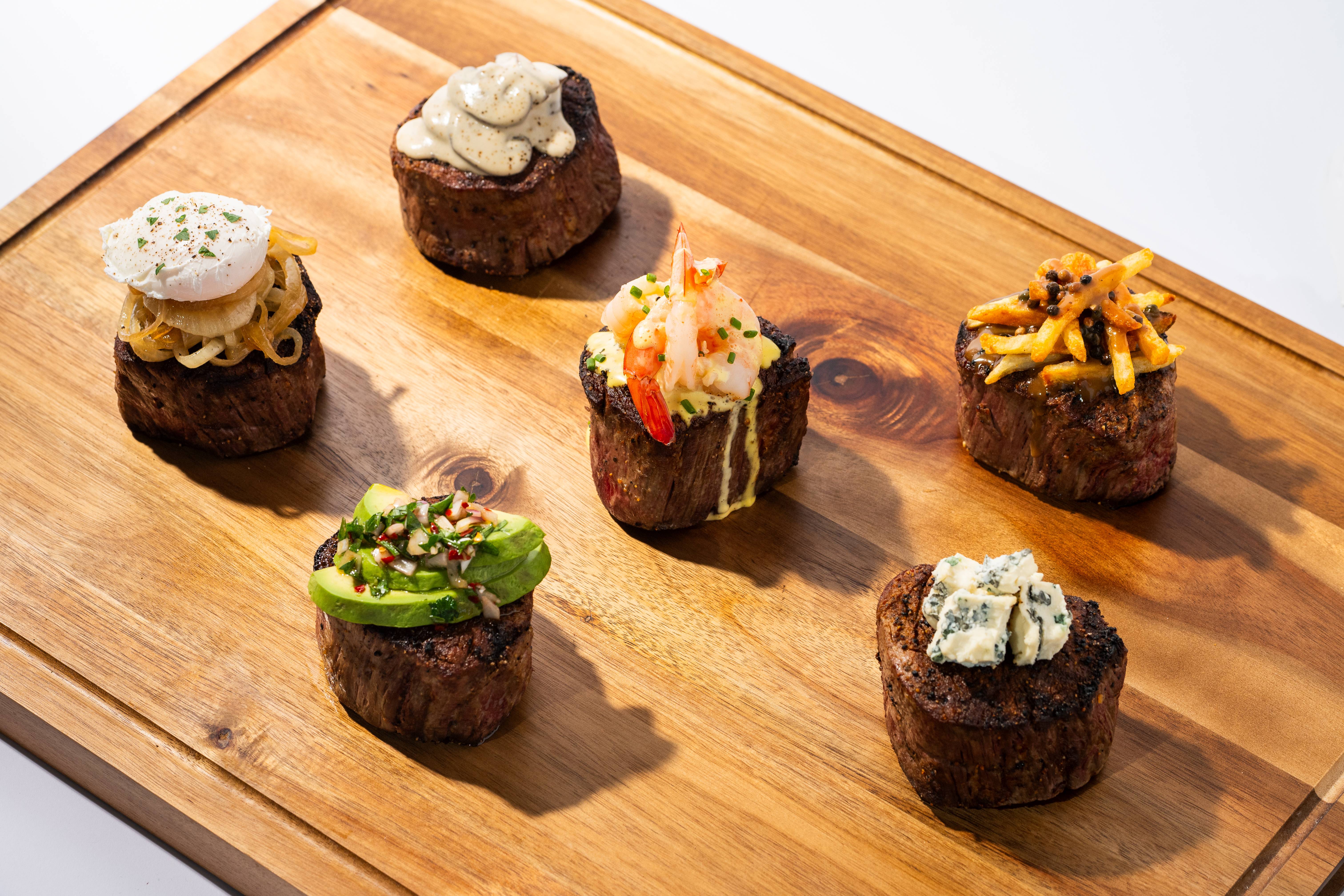 Small cuts of filet mignon on a wooden board with five toppings, including shrimp, avocado, blue cheese, and crispy fries.