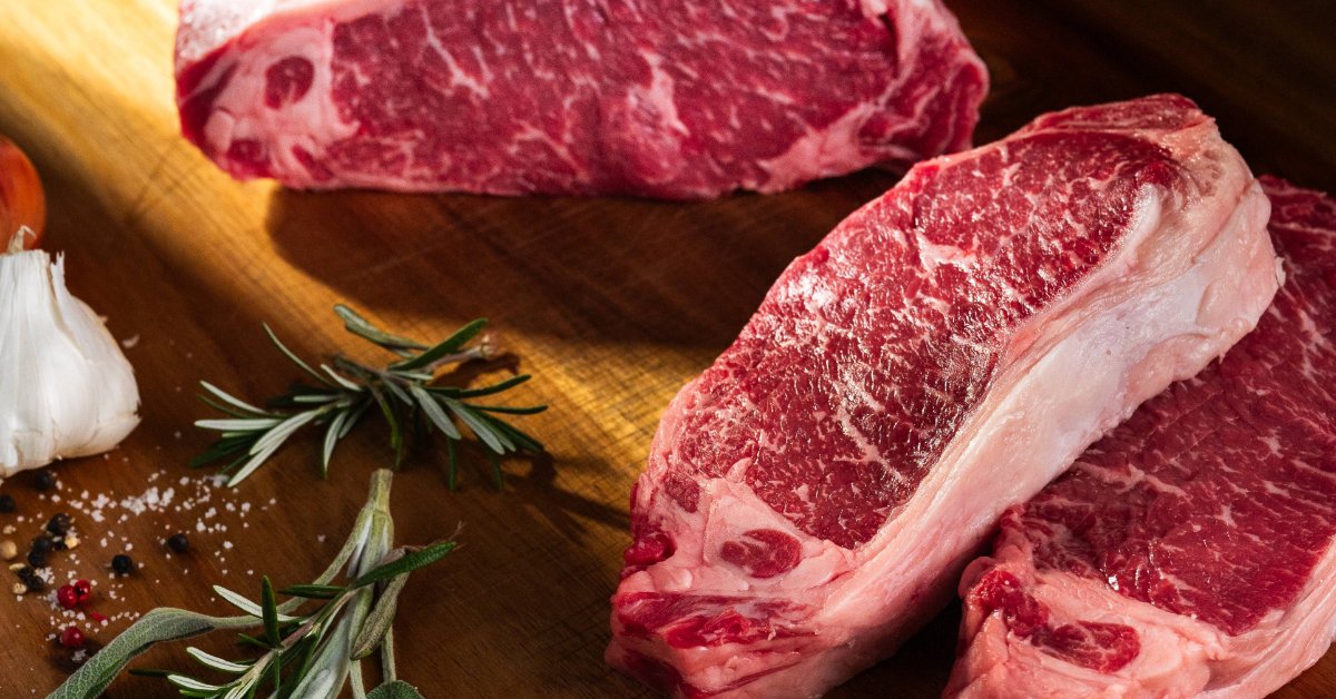How To Cook the Perfect Strip Steak in a Cast-Iron Skillet Raw New York Strip steaks and a whole beef roast rest on a wood board styled with fresh herbs and garlic cloves nearby.