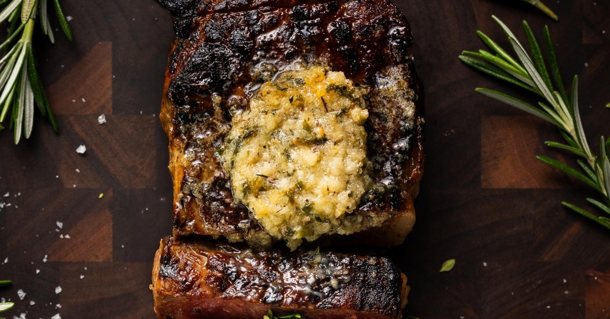 How To Cook the Perfect Strip Steak in a Cast-Iron Skillet A sliced steak with a deep char and pink center, topped with melted herb butter and surrounded by fresh rosemary.