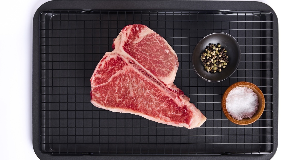 An uncooked porterhouse steak with visible marbling rests on a wire rack next to salt and mixed peppercorns. An uncooked porterhouse steak with visible marbling rests on a wire rack next to salt and mixed peppercorns.