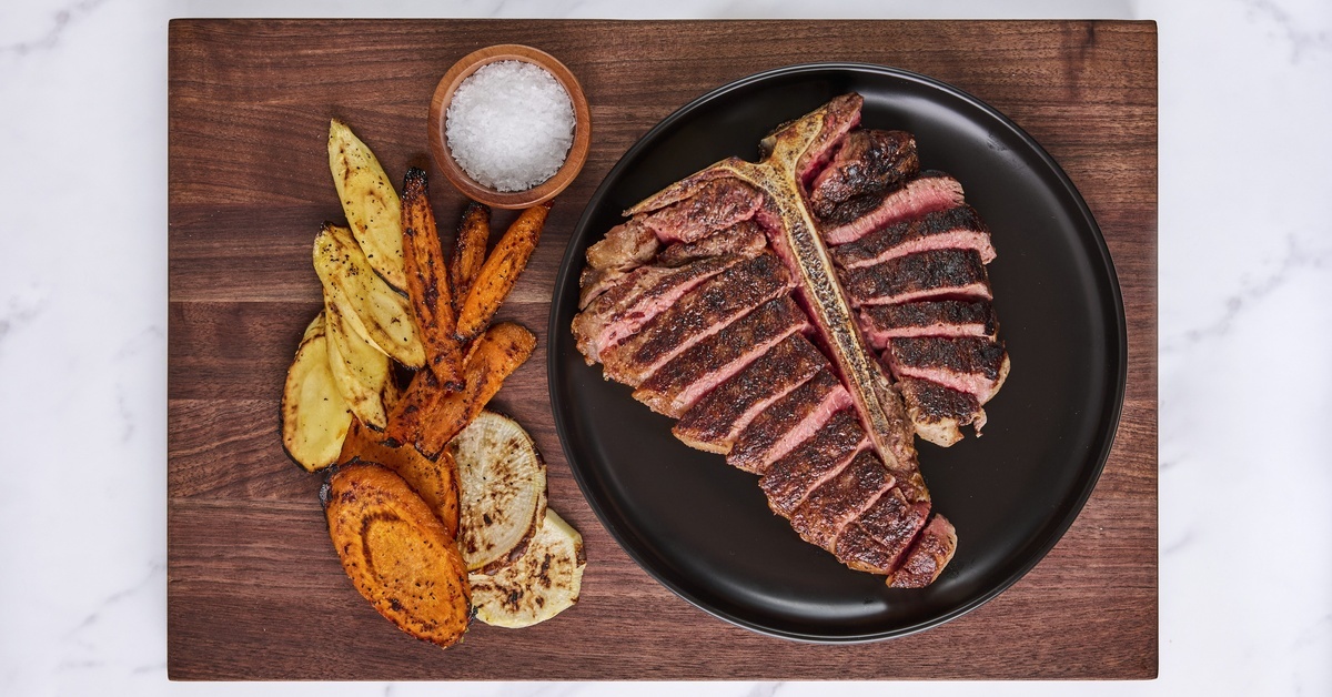Overhead shot of a sliced steak on a black plate next to grilled vegetables and a small dish of salt on walnut wood. Overhead shot of a sliced steak on a black plate next to grilled vegetables and a small dish of salt on walnut wood.