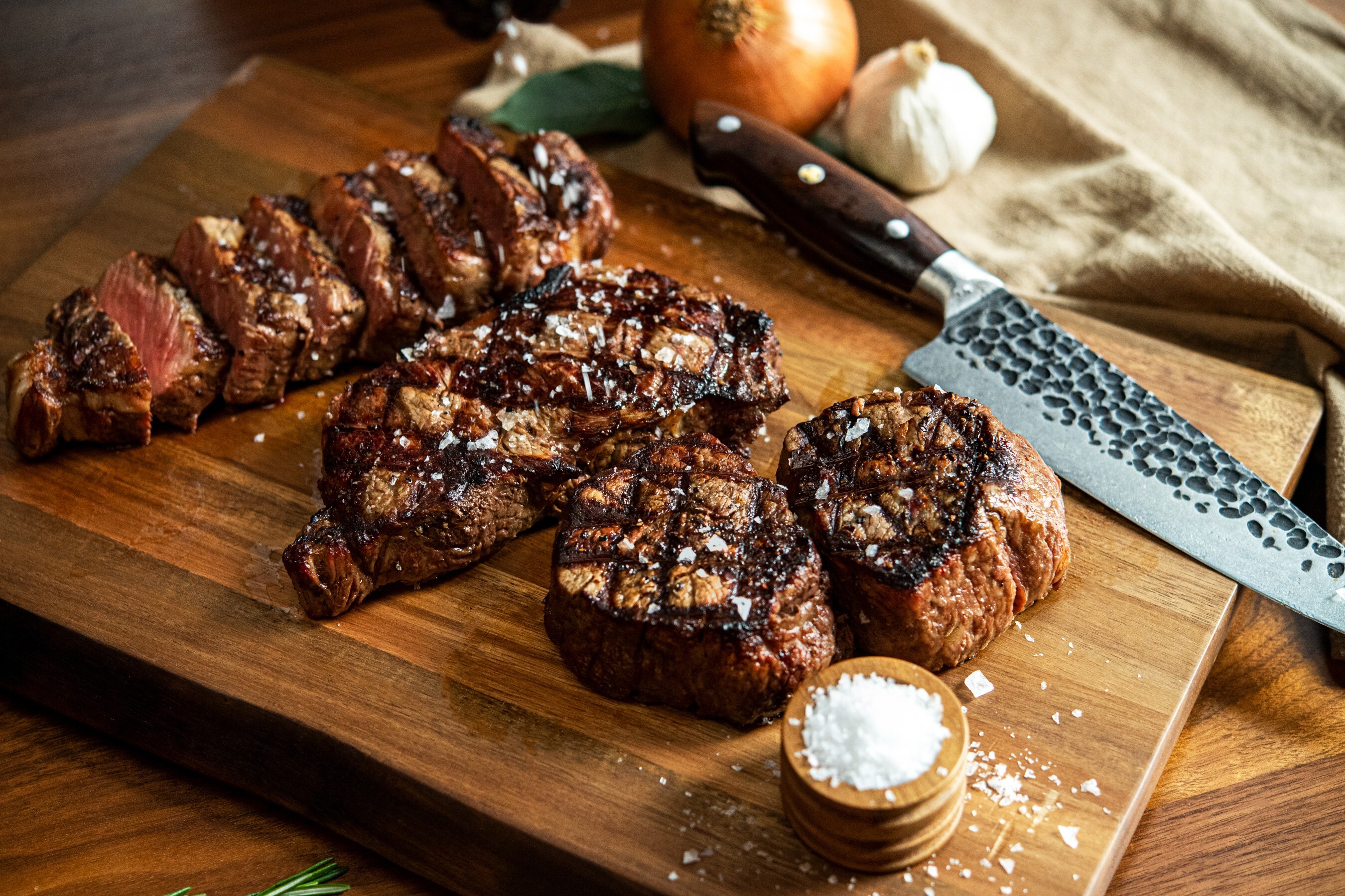 Grilled filet steaks, one sliced to show a rosy pink center, resting on a wooden board with sea salt and a chef knife.