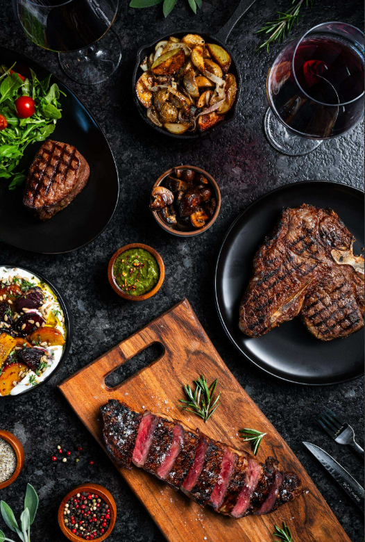 An aerial view shows a tablescape with cooked steak, glasses of wine, chimichurri sauce, peppercorns, and silverware.