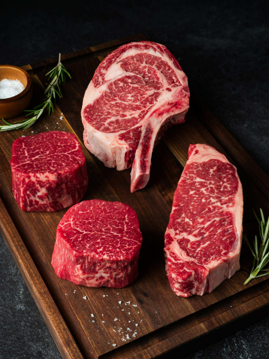 An uncooked bone-in ribeye, strip steak, and two Filet Mignons sit on a wooden cutting board by salt and fresh rosemary.