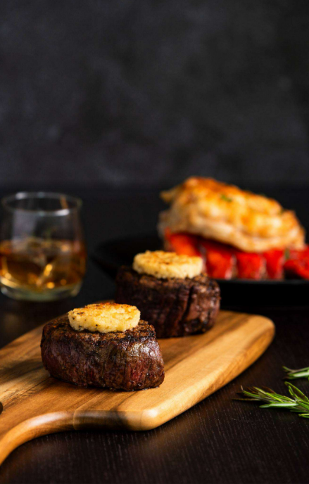 Two cooked filet mignon steaks with a compound butter in the center sit on a wooden cutting board.