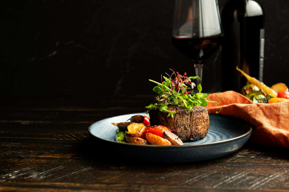 A cooked filet mignon with a garnish of greens sits on a round plate beside roasted potatoes. A glass of wine is behind it.