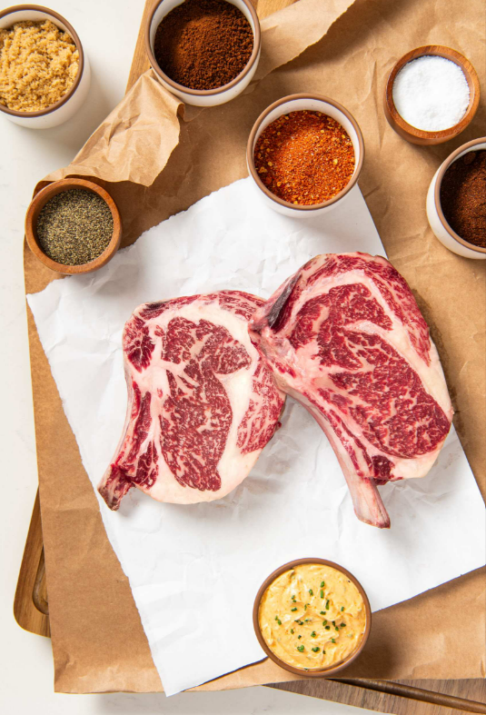 Anatomy of an Unforgettable Ribeye Steak Two large, uncooked bone-in ribeye steaks sit on a white parchment. Small bowls of various spices surround the steaks.