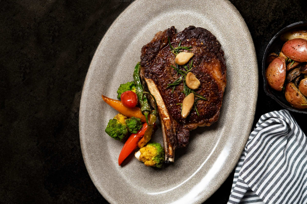Anatomy of an Unforgettable Ribeye Steak A cooked bone-in ribeye steak sits on a speckled gray plate next to a pile of roasted vegetables.
