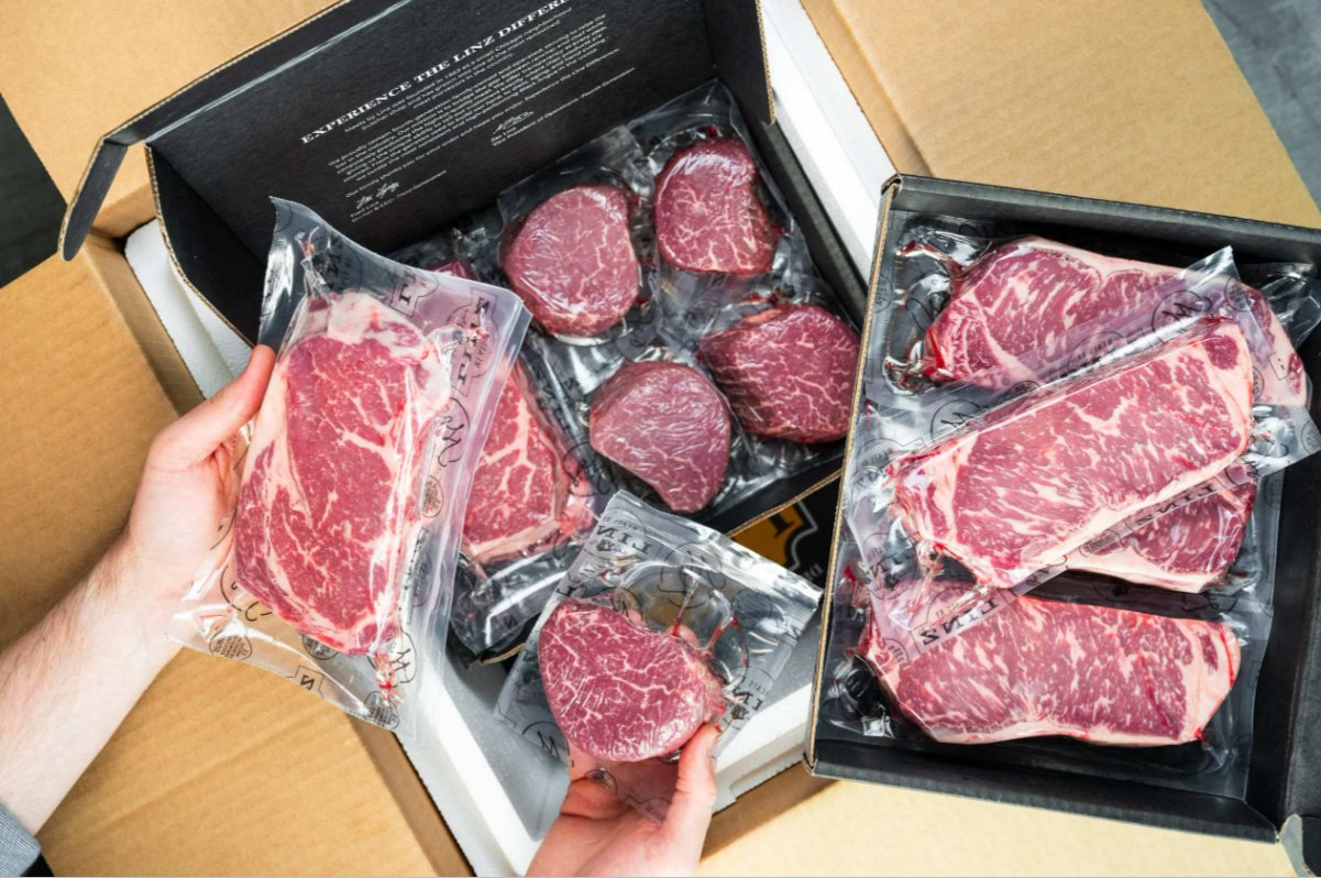 Why Sourcing 100% Black Angus Beef Makes a Difference in Premium Steaks A person is holding two different packaged steaks above a cardboard box full of other packaged steaks.