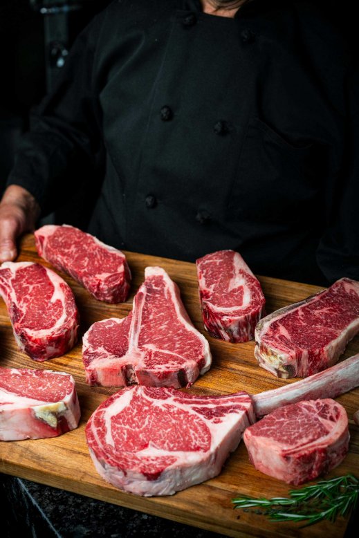 Dry-Aged Beef Timeline: A Timeline of Flavor at Every Stage Several different cuts of steak sit on a wooden cutting board with a person holding the board. The steaks are uncooked.