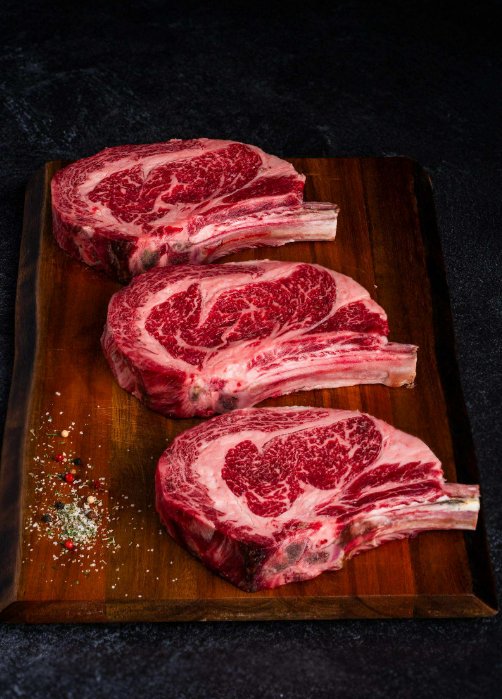 Dry-Aged Beef Timeline: A Timeline of Flavor at Every Stage Three uncooked, bone-in steaks sit on a wooden cutting board. A pile of colorful peppercorns sits in the corner of the board.