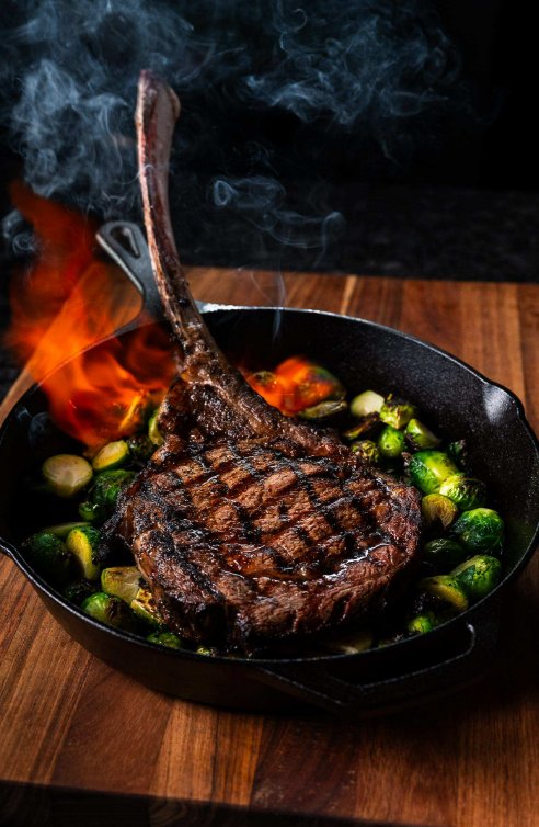 Tomahawk vs Cowboy Ribeye: What Sets Them Apart A bone-in steak sits in a cast-iron skillet with roasted Brussels sprouts and flames. The skillet sits on a cutting board.