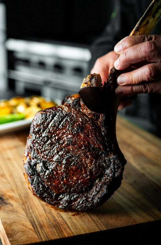 Tomahawk vs Cowboy Ribeye: What Sets Them Apart A close-up view shows a person holding the bone of a cooked steak while using a knife to cut the steak.