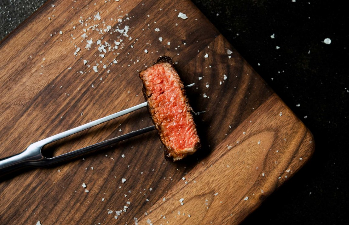 What To Know Before You Order Steaks Online A close-up view shows a slice of cooked steak skewered by a meat fork and resting on a wooden cutting surface.