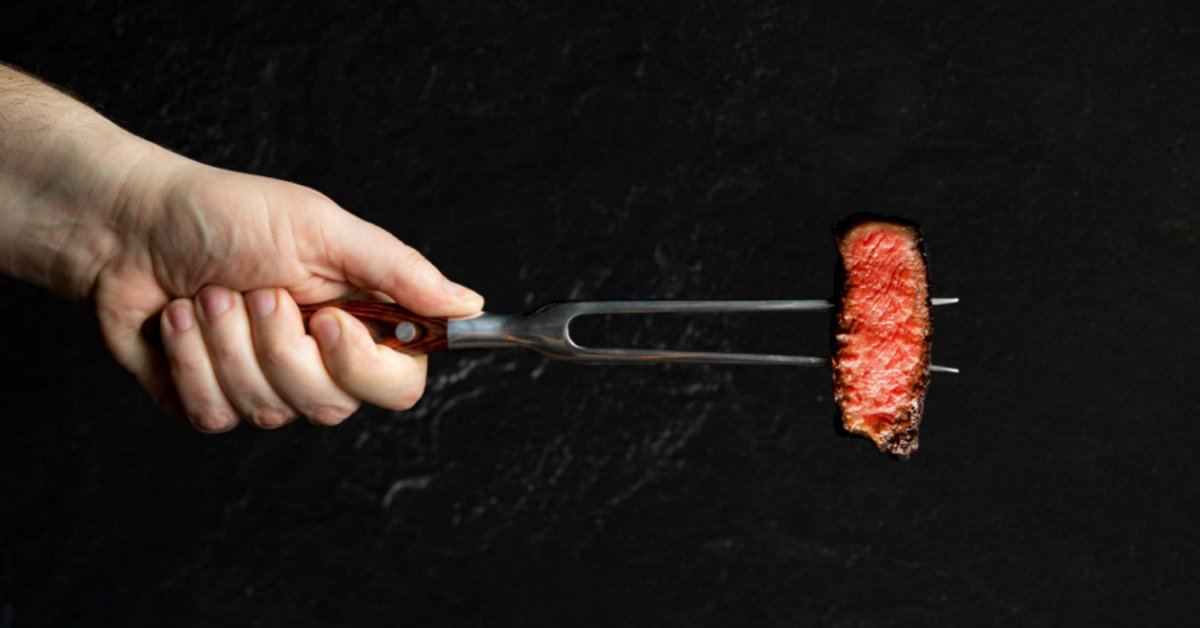 How To Get Steakhouse Results With Filet Mignon A person's hand holds a stainless steel carving fork that contains a piece of cooked steak. The background is black.