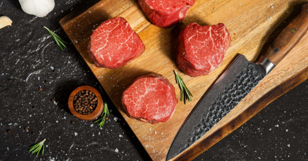 How To Get Steakhouse Results With Filet Mignon Four uncooked filet mignon steaks sit on a wooden cutting board next to a chef's knife and a sprig of fresh rosemary.