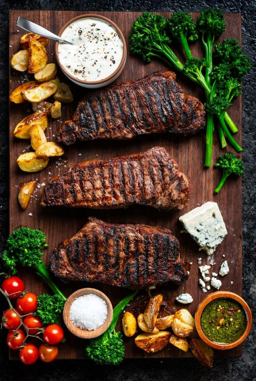 What Separates a Great New York Strip Steak Three cooked strip steaks sit on a wooden cutting board beside bowls of sauce, cherry tomatoes, cooked potatoes, and broccoli.