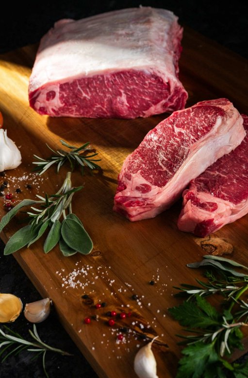 What Separates a Great New York Strip Steak Two raw strip steaks sit on a wooden cutting board next to a chunk of raw beef, salt, peppercorns, sage, garlic, and rosemary.