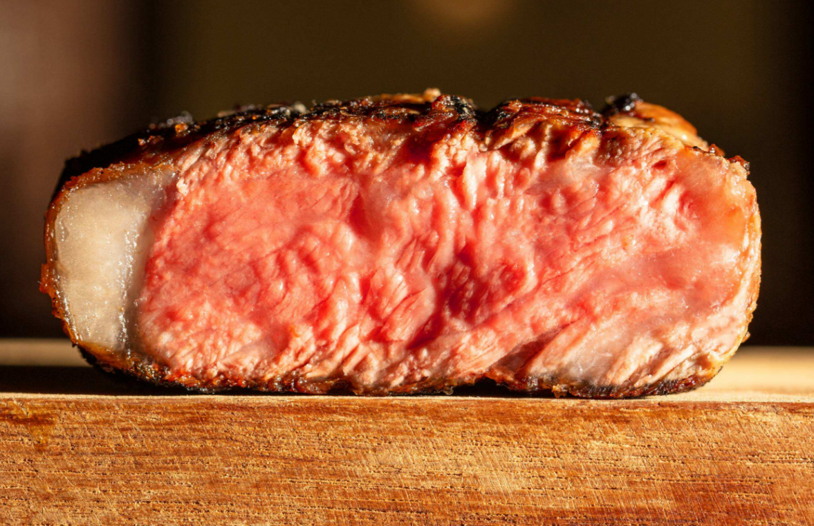 A close-up view shows a slice of a cooked piece of steak. The steak slice sits on a wooden cutting board.