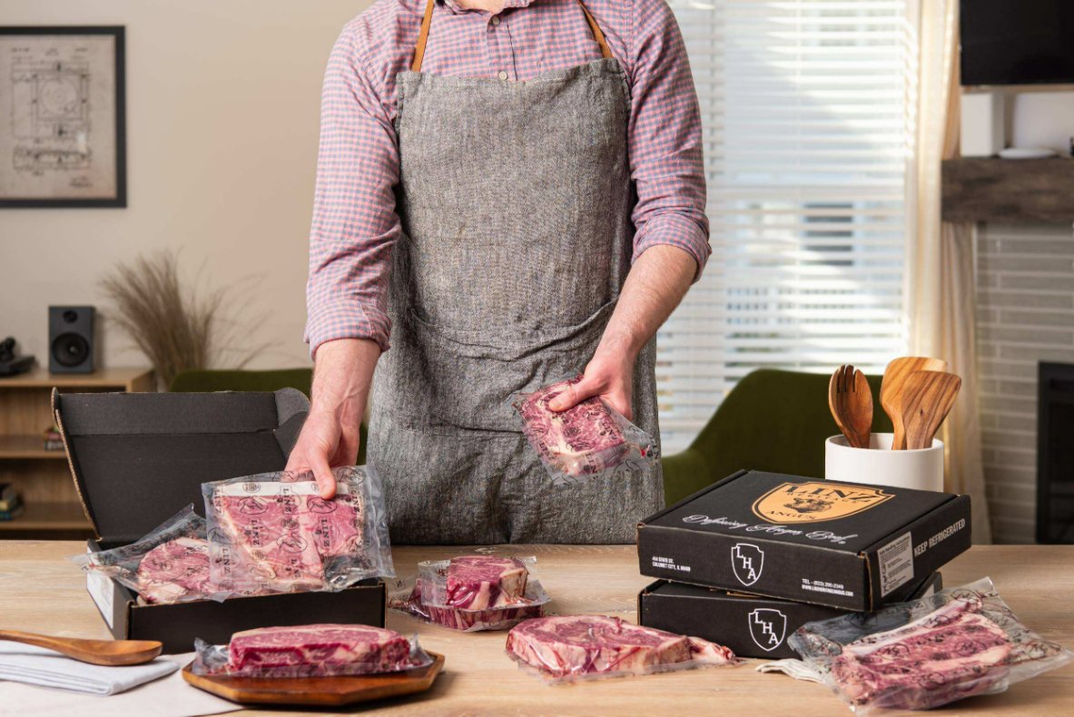How Online Steak Delivery Actually Works A man wearing a gray apron is grabbing packaged steaks from a black box and placing them onto a wooden table.