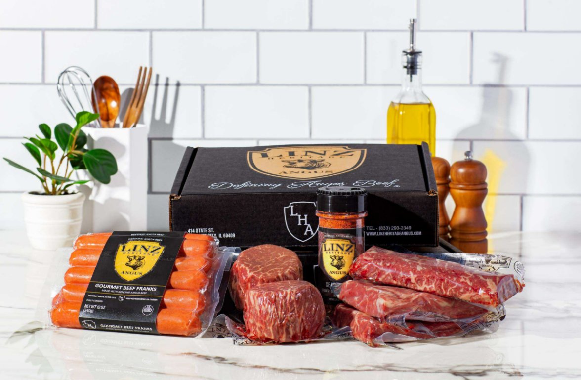 How Online Steak Delivery Actually Works Packaged beef franks, filet mignon, strip steaks, and seasoning mix sit in front of a black box on a marbled surface.