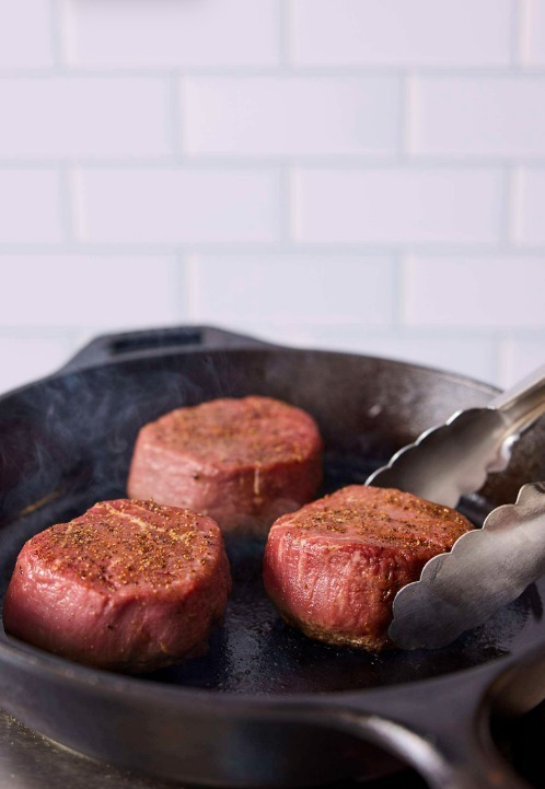 Three filet mignons are cooking in a cast iron skillet. A pair of metal cooking tongs is flipping one of the filets.