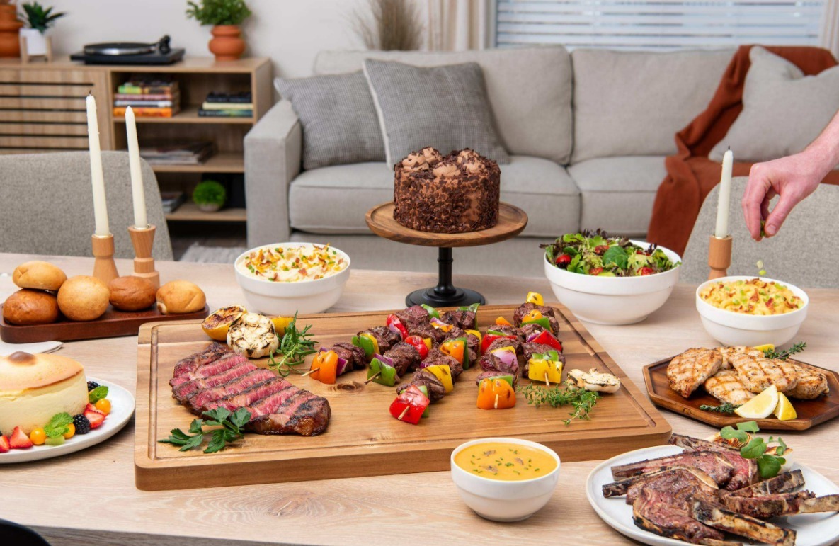 A table features a wooden cutting board with cooked steak, serving bowls with sides, and a pair of candlesticks.