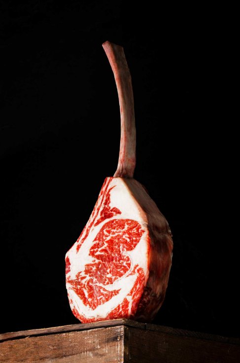 Tomahawk Steak Guide for First-Time Buyers A raw tomahawk steak sits on a wooden surface on its side with the bone side up in the air. The background is black.