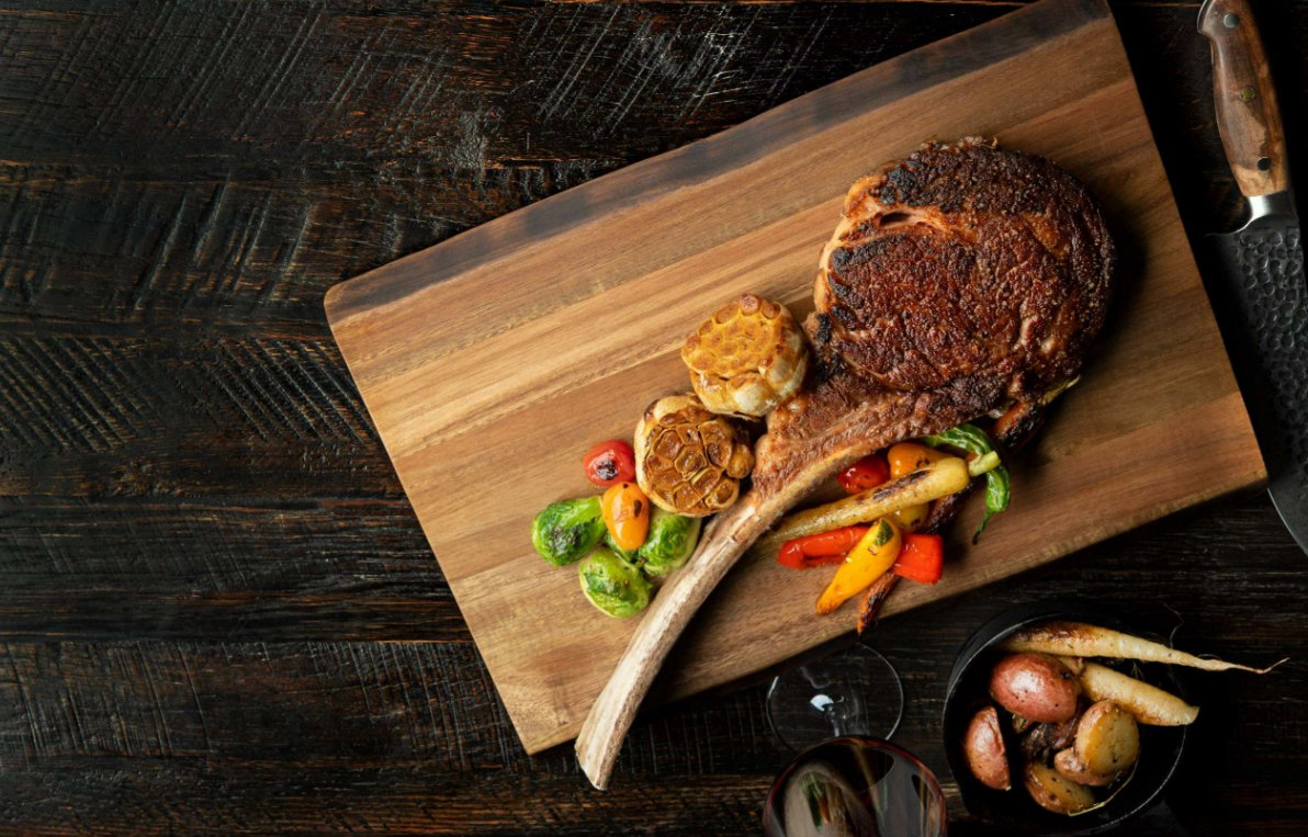 Tomahawk Steak Guide for First-Time Buyers A cooked tomahawk steak sits on a wooden cutting board next to two roasted heads of garlic and roasted vegetables.