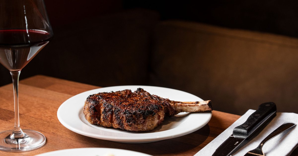 Tips for Choosing Wine Pairings for Ribeye Steak A bone-in ribeye steak served on a white plate next to a glass of red wine on a wooden restaurant table.