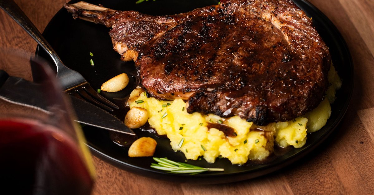 Tips for Choosing Wine Pairings for Ribeye Steak A bone-in ribeye steak served over mashed potatoes on a black plate, accompanied by a glass of red wine.