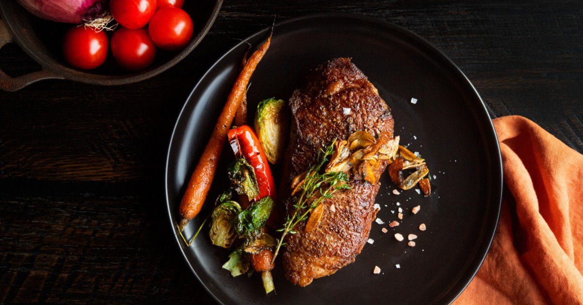 A seared steak on a black plate with roasted vegetables, coarse salt, herbs, tomatoes, and an orange napkin. A seared steak on a black plate with roasted vegetables, coarse salt, herbs, tomatoes, and an orange napkin.