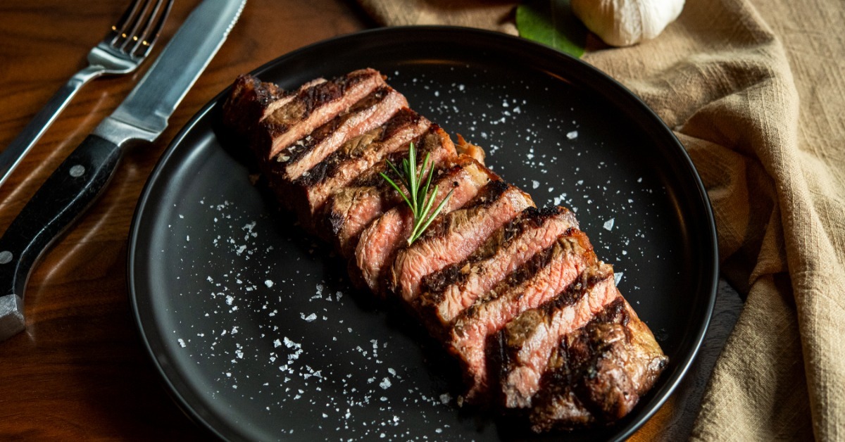 Sliced grilled steak on a black plate with coarse salt, rosemary, a fork, a knife, garlic, and a beige cloth. Sliced grilled steak on a black plate with coarse salt, rosemary, a fork, a knife, garlic, and a beige cloth.